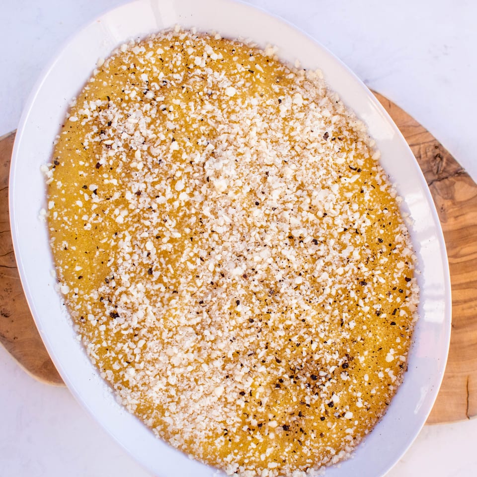 A white oval dish filled with golden polenta, topped with white cheese and black pepper.
