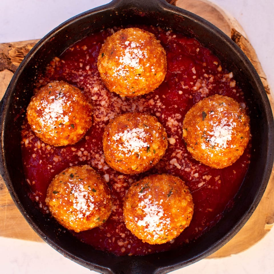 Six golden-brown arancini, sprinkled with Parmesan, rest in a rich red tomato sauce in a cast-iron pan.