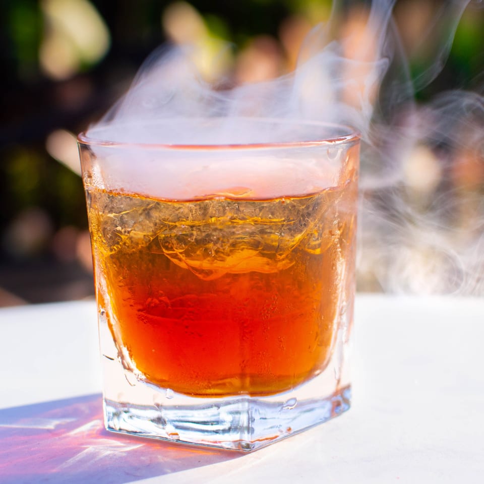 A PINO Italian restaurant cocktail, amber-colored with ice and dramatic smoke, sits on a white surface.
